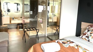 Une chambre d’hôtel moderne dotée d’une douche à parois de verre, d’un lit avec une couvre-lit à motifs, d’un petit coin salon et d’un grand miroir.
