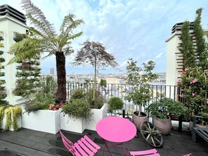 Une terrasse sur le toit offrant une vue sur la ville, agrémentée de plantes en pot, d’une table rose et d’un jardinière en forme de roue de vélo.