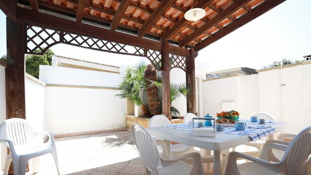 A covered patio with a table set for dining, surrounded by white plastic chairs, and a white wall in the background. - Villetta Anna Torre Suda (Racale)