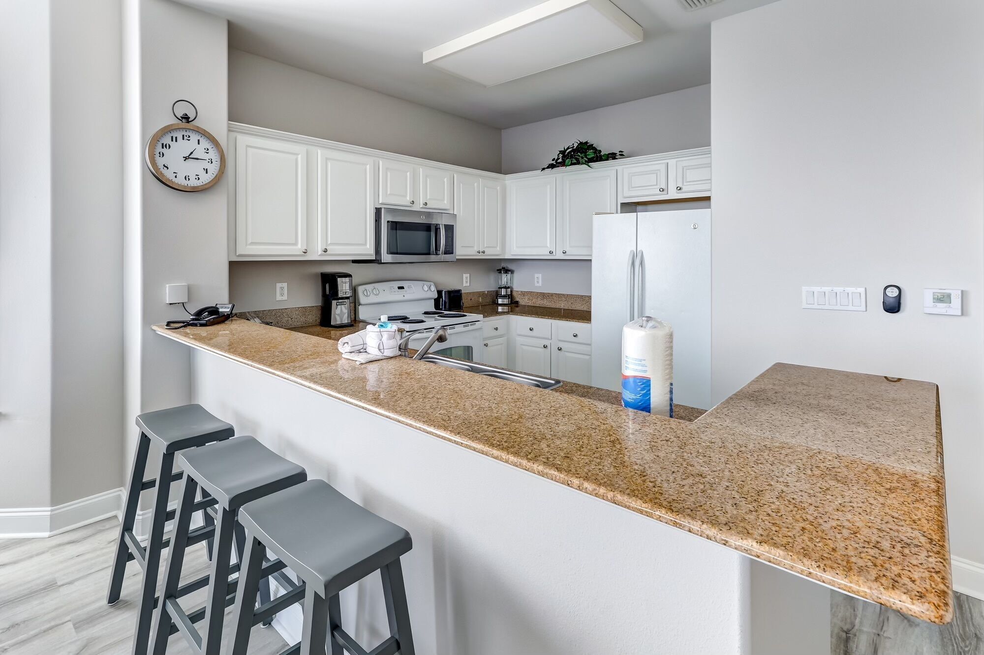 Una cocina moderna con armarios blancos, encimera de granito, microondas, refrigerador y cafetera. Hay tres taburetes de bar y un reloj en la pared.