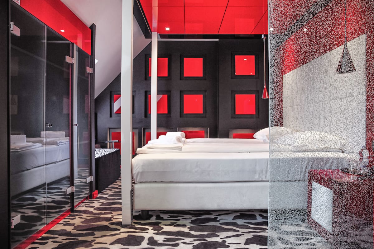 A modern hotel room with a glass partition, a bed with white linens, a patterned carpet, and red accents on the walls and ceiling.