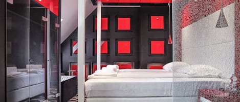 Una habitación de hotel moderna con una pared divisoria de vidrio, una cama con ropa blanca, una alfombra estampada y detalles rojos en las paredes y el techo.