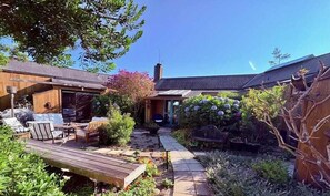Eine Holzdielen-Terrasse mit Gartenmöbeln, ein Garten mit verschiedenen Pflanzen und ein Gebäude mit einem geneigten Dach.