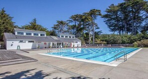 Piscina com área de deck de madeira, um edifício com telhado inclinado e árvores ao fundo.
