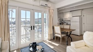 Un salon moderne doté d’une grande porte-fenêtre coulissante donnant sur un balcon, d’un coin repas avec une table et des chaises en bois, et d’une cuisine compacte avec des meubles et des appareils électroménagers blancs.