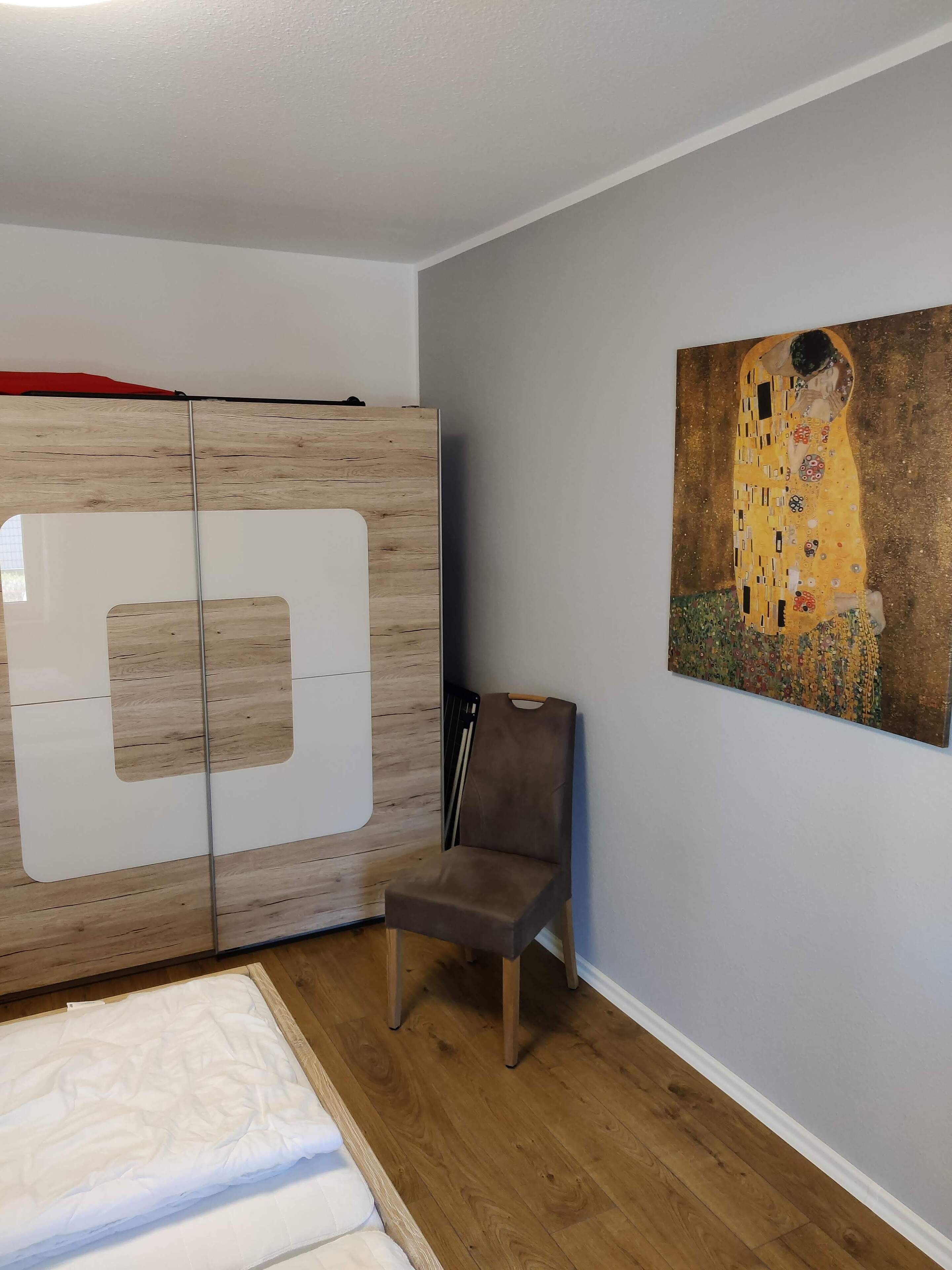 Ein Schlafzimmer mit einem Holzschrank, einem grauen Sessel und einem Bild an der Wand.