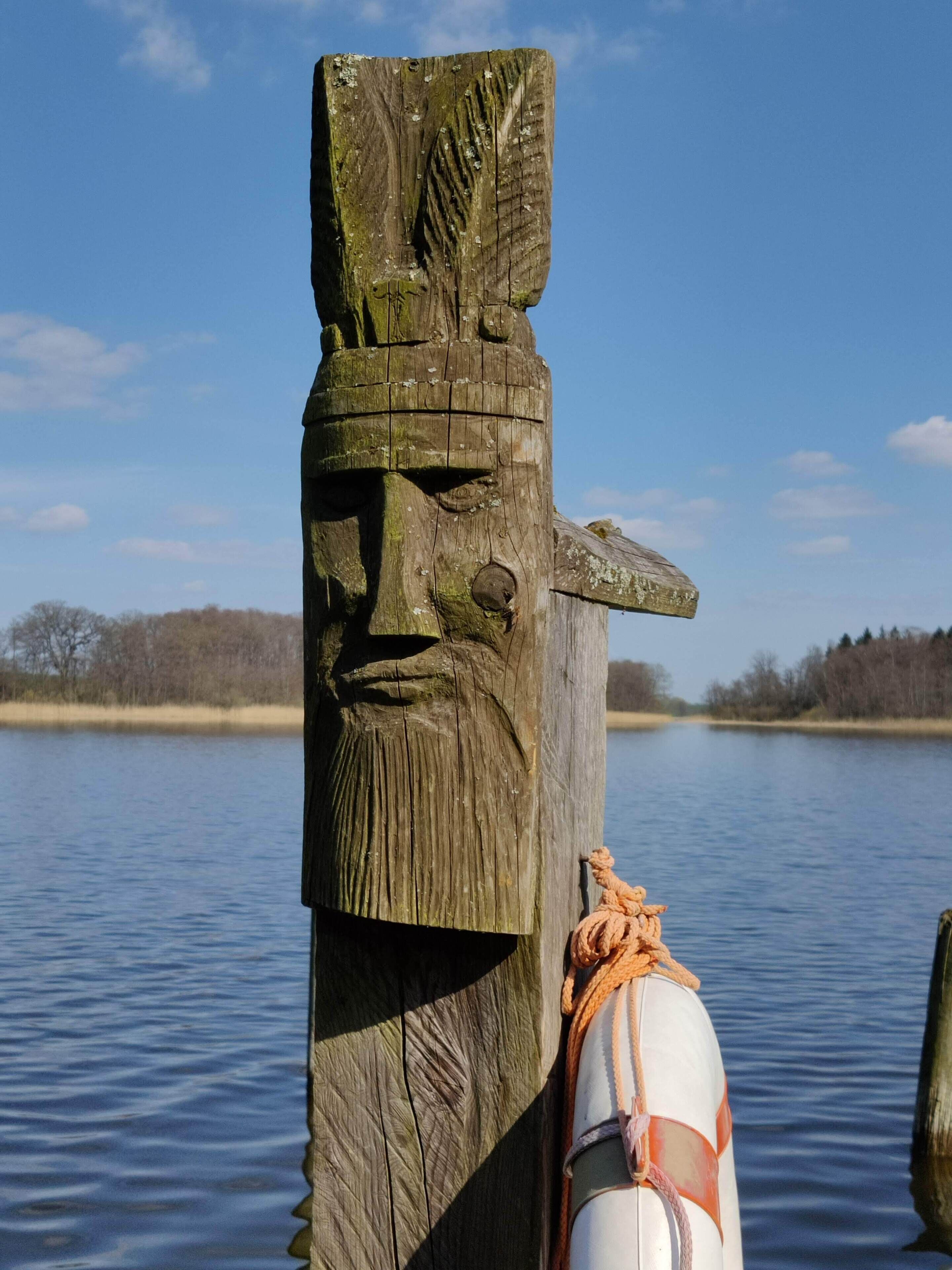 Een houten paal met een uitgehouwen gezicht, omwikkeld met touw, en een boei in het water.