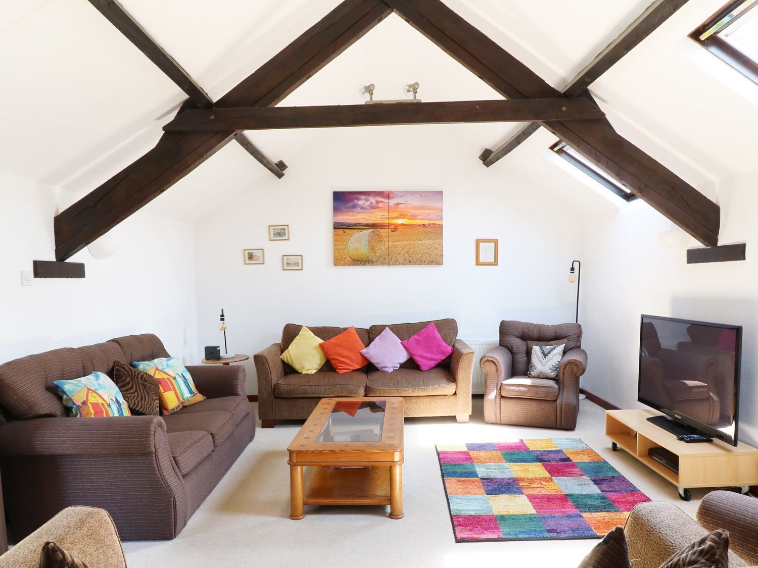 A spacious living room with a high ceiling, exposed wooden beams, a large window, a sofa set, a coffee table, a television, and a colorful rug.