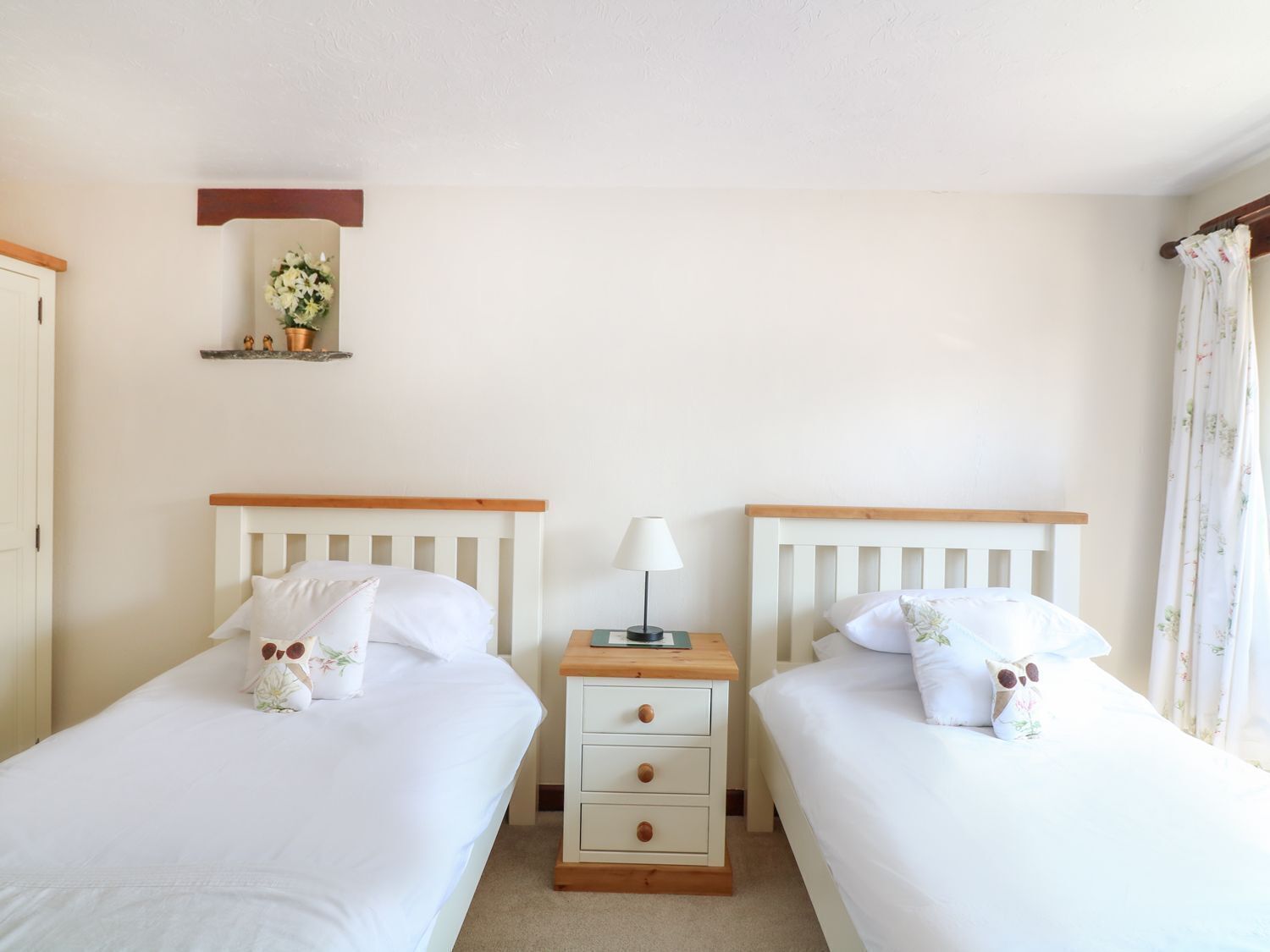 Een tweepersoonskamer met twee bedden, een nachtkastje met een lamp, een klein plankje met bloemen en een deur aan de linkerkant.