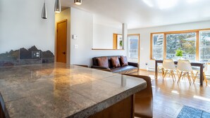 Een moderne keuken met een groot eiland, een eethoek met een houten tafel en stoelen, en een zithoek met een bank en kussens.