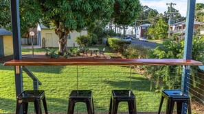 A patio with a wooden table and three black stools, overlooking a well-maintained lawn and a residential street. - Torquay Treasure - Located in beautiful Hervey Bay less than 100m walk to the beach (Torquay)