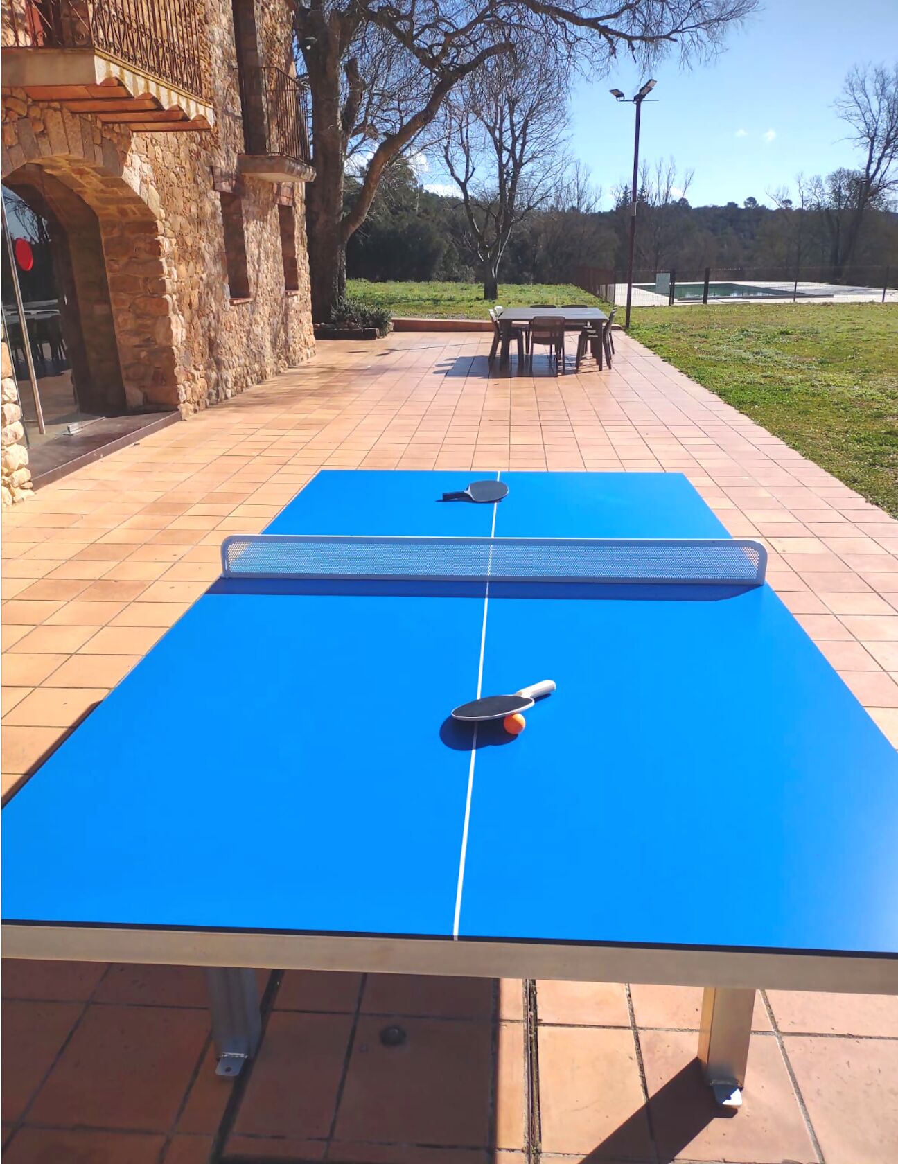 An outdoor ping pong table with a blue surface and two paddles, set on a tiled patio with stone walls and a grassy area in the background.