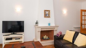 A living room with a flat-screen TV on a white stand, a fireplace with a mantelpiece, a black leather sofa with cushions, and a wooden coffee table. - Burgo D. Sebastião 27B (Óbidos)