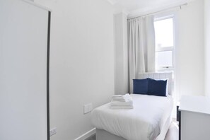 Un dormitorio minimalista con cama, ventana con cortinas, radiador y mesita de noche.