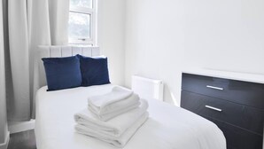 Ein ordentlich bezogenes Bett mit weißen Leinen und blauen Kissen, ein weißer Heizkörper und eine dunkle Holzkommode mit Schubladen.