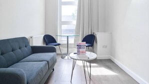 Ein modernes Wohnzimmer mit einem grauen Sofa, zwei blauen Sesseln, einem Glastisch und Blick ins Freie durch ein Fenster mit weißen Vorhängen.
