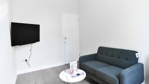 Ein Wohnzimmer mit flachbildfernseher an der Wand, einem grauen Sofa, einem kleinen weißen Couchtisch und einer weißen Tür.
