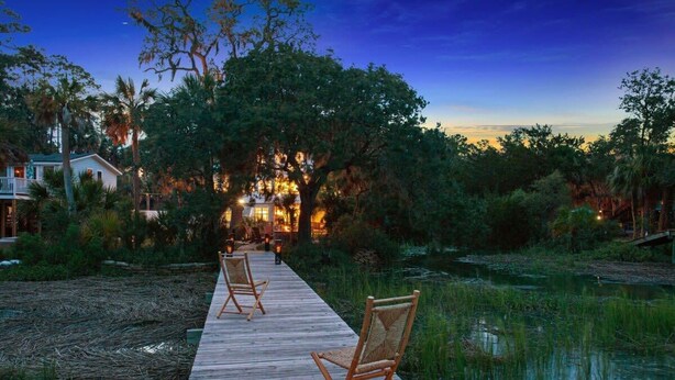 A wooden deck with chairs leading to a building with warm lighting, surrounded by trees and a body of water. - Large Peaceful Home | Hot Tub, Dock Near Beach (Savannah)