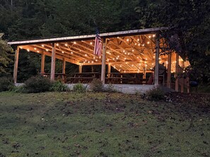 A wooden pavilion with string lights and an American flag, surrounded by greenery. - Beautiful 4 unit lodge w/ Hot tub, WiFi, fire pits &  pavilion area with grills (Stanton)