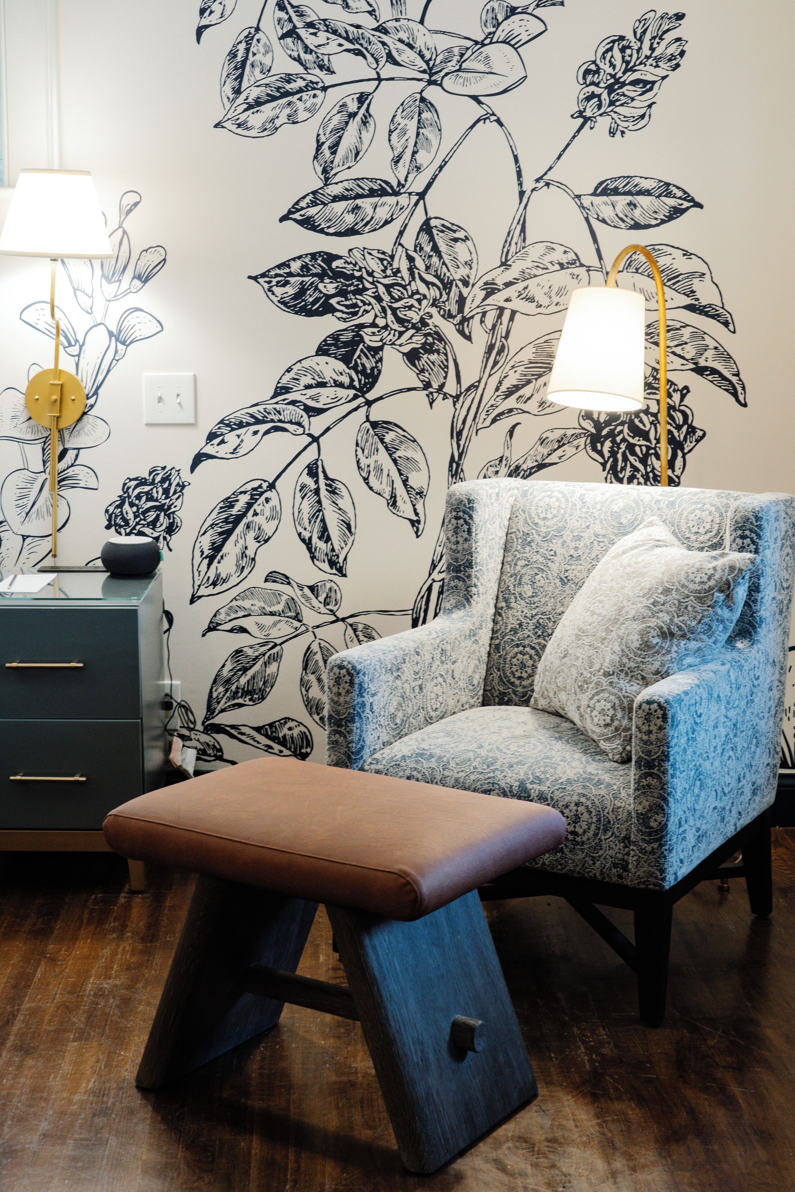 A room with a patterned wall, a chair with a cushion, a bench, a lamp, and a desk with drawers.