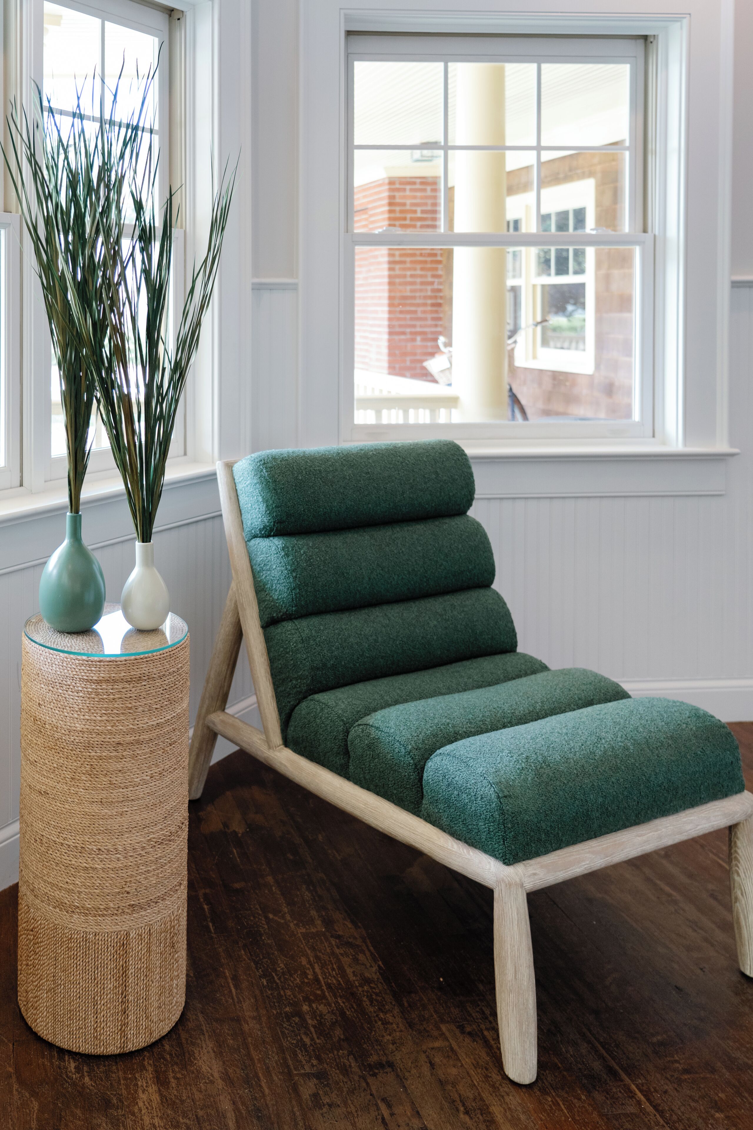 Una habitación con una silla verde acolchada, una mesita auxiliar trenzada y un jarrón alto con plantas verdes.