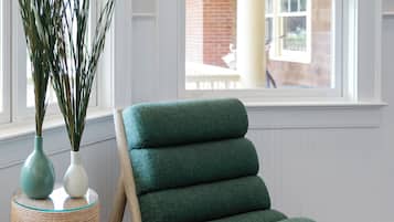 Una habitación con una silla verde acolchada, una mesita auxiliar trenzada y un jarrón alto con plantas verdes.