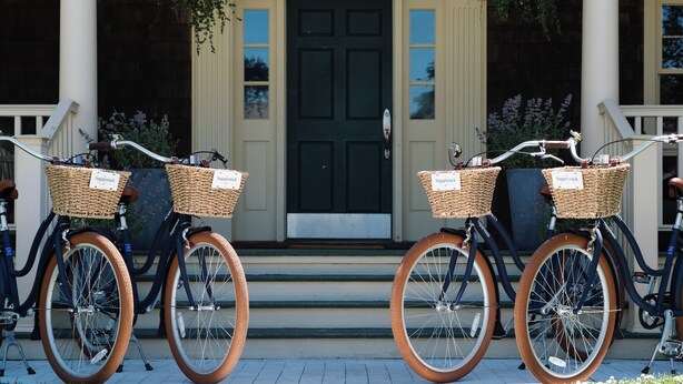 Deux vélos équipés de paniers en osier portant l’inscription « Sophie & Co. » sont garés devant un immeuble dont la porte est sombre et qui est orné de colonnes blanches.