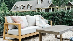 Un espace extérieur aménagé avec des meubles en bois, comprenant un canapé et une table, entouré de gravier et de verdure, avec une maison en arrière-plan.