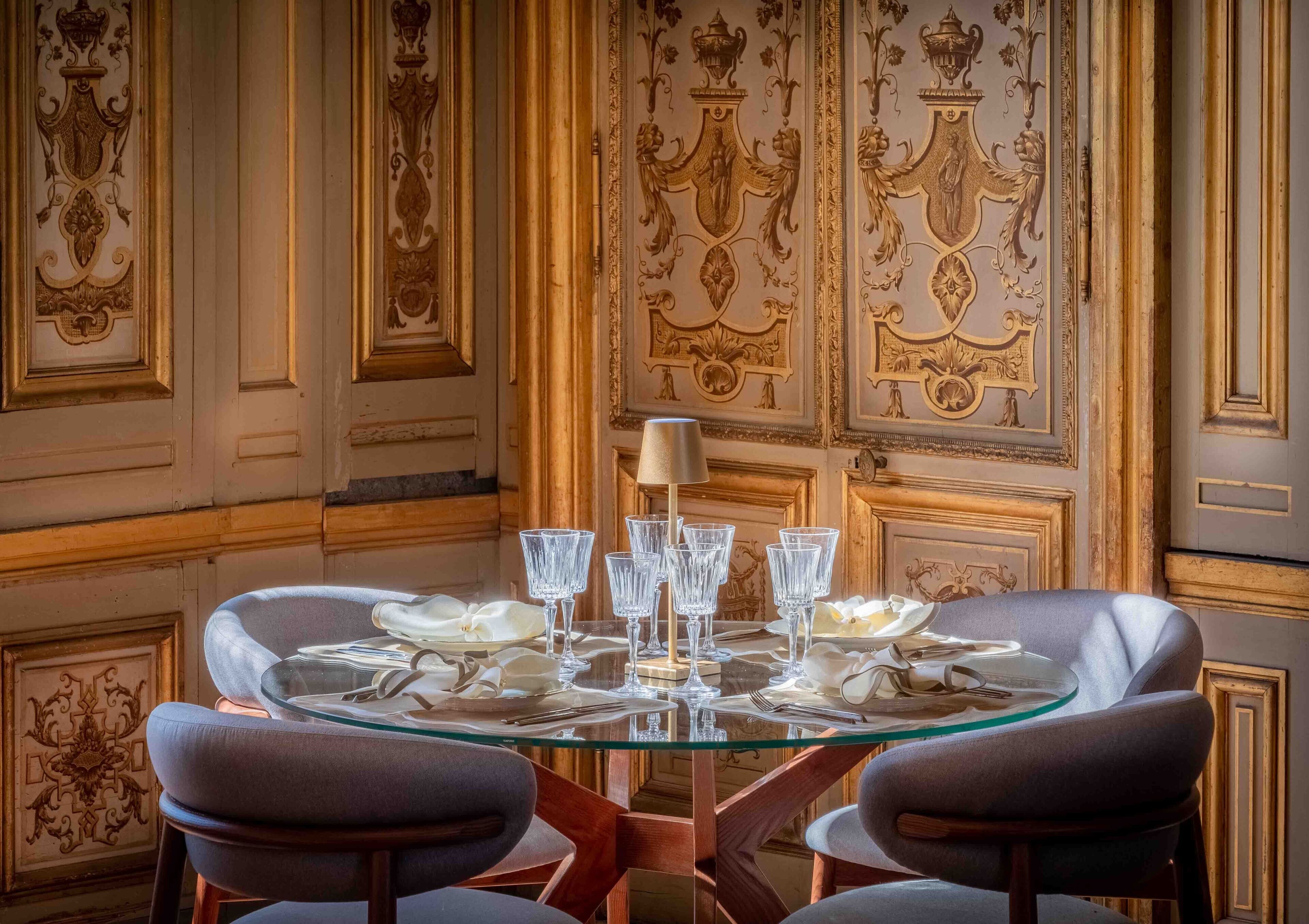 A dining table set with crystal glassware and plates, surrounded by upholstered chairs, in a room with ornate wall decorations.