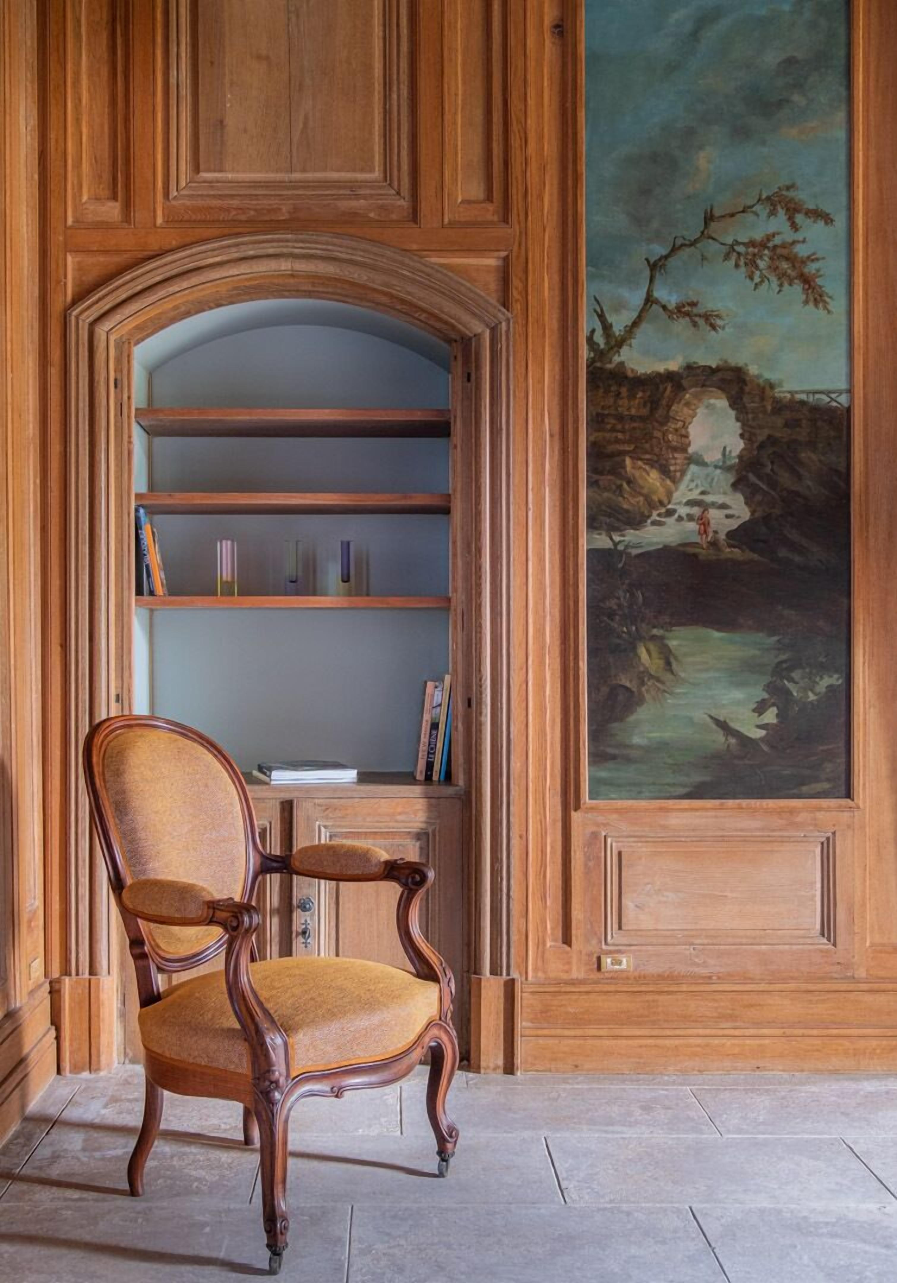 A wooden bookshelf with a chair in front, a painting on the wall, and a stone floor.