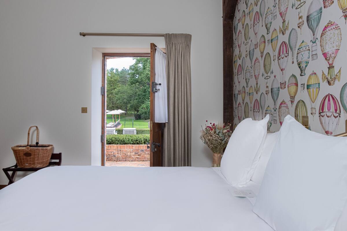 Een slaapkamer met een groot bed, een rieten mand, een vaas met bloemen en uitzicht op een tuin door een geopende deur.