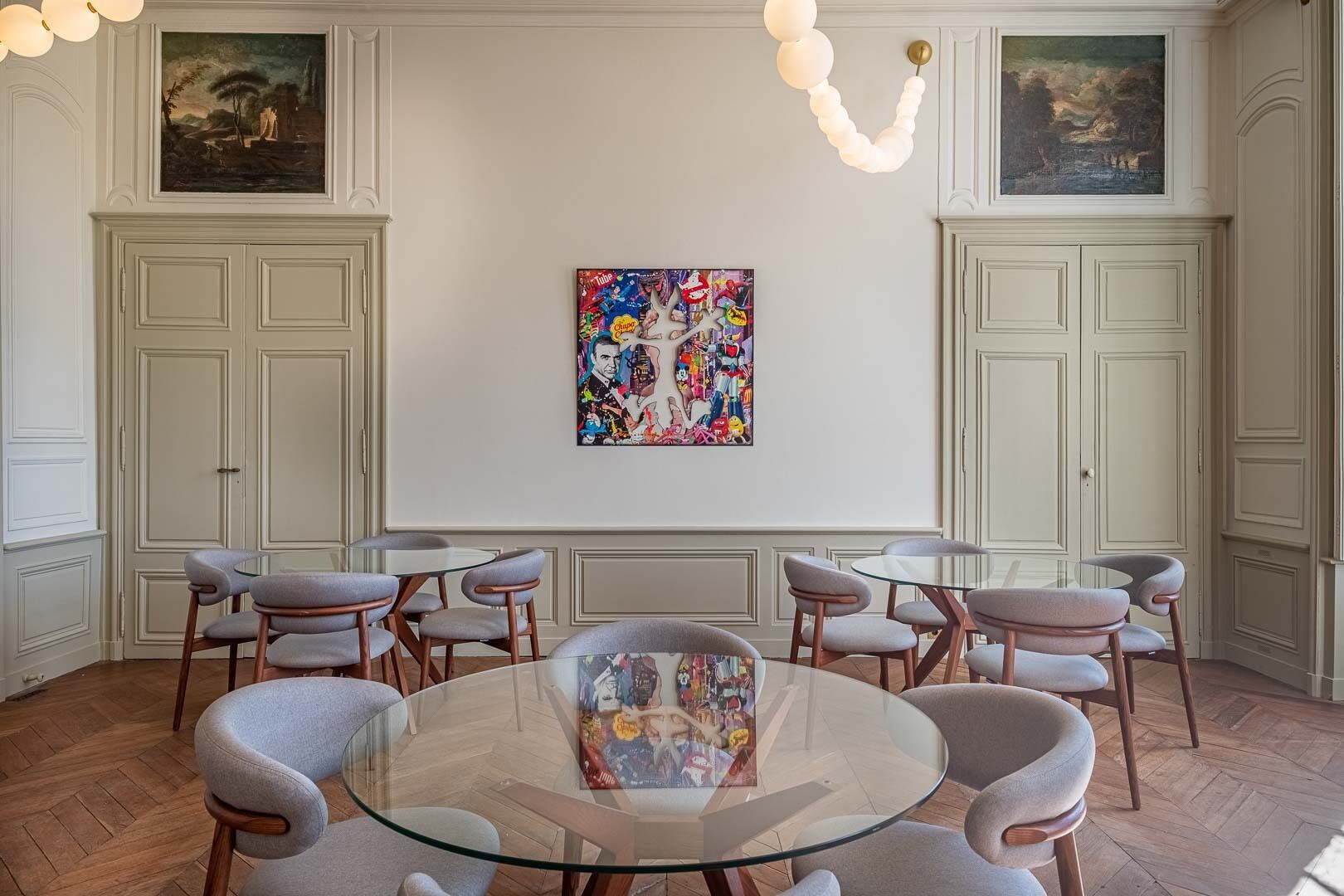 A dining area with a glass table surrounded by chairs, a colorful abstract painting on the wall, and classic white paneling.