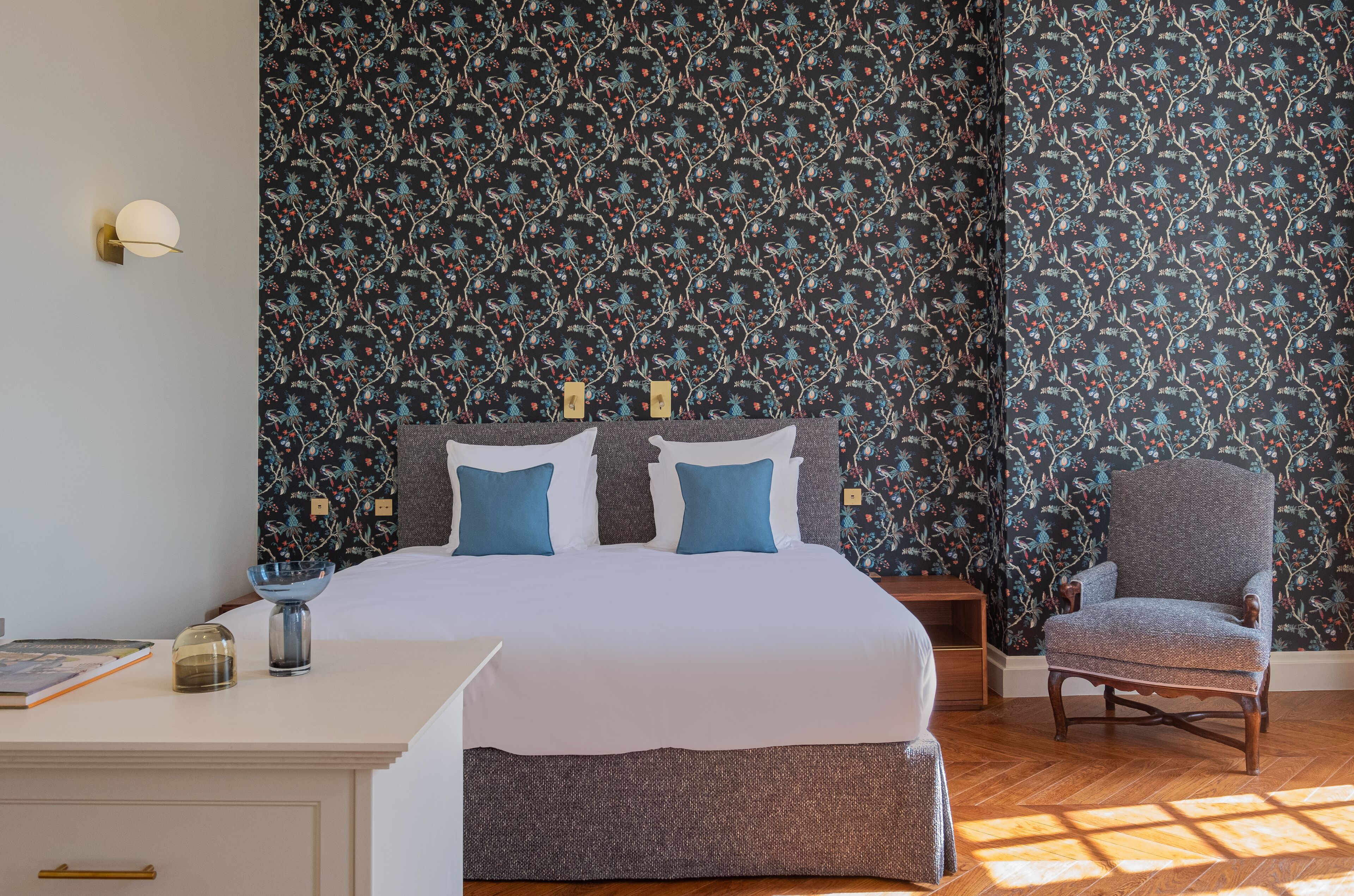 Une chambre à coucher comprenant un lit, deux tables de chevet, une chaise et un mur orné d’un motif floral.