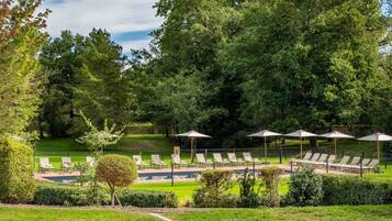 Een zwembadterras met ligbedden en parasols, omgeven door weelderige begroeiing en bomen.