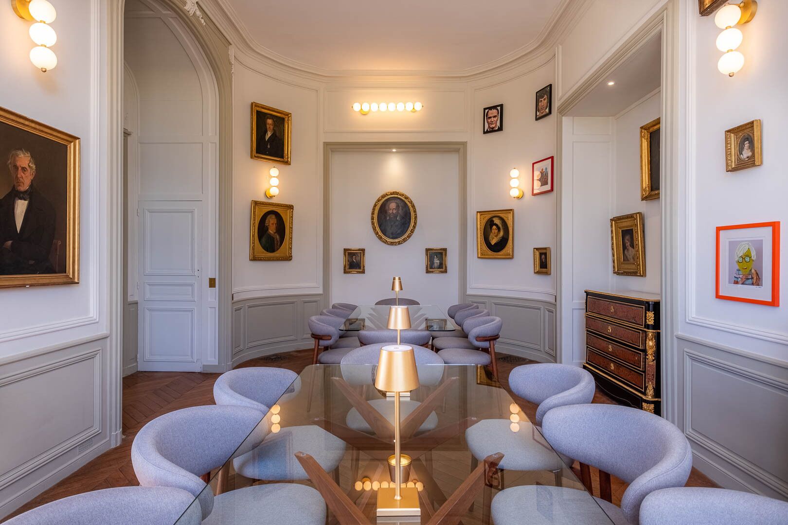 A formal meeting room with a glass table, grey upholstered chairs, and framed portraits on the walls.