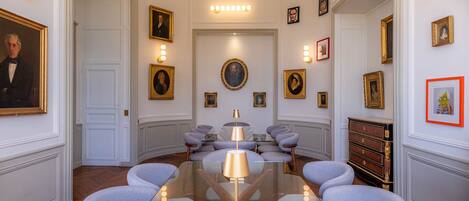 A formal meeting room with a glass table, grey upholstered chairs, and framed portraits on the walls.