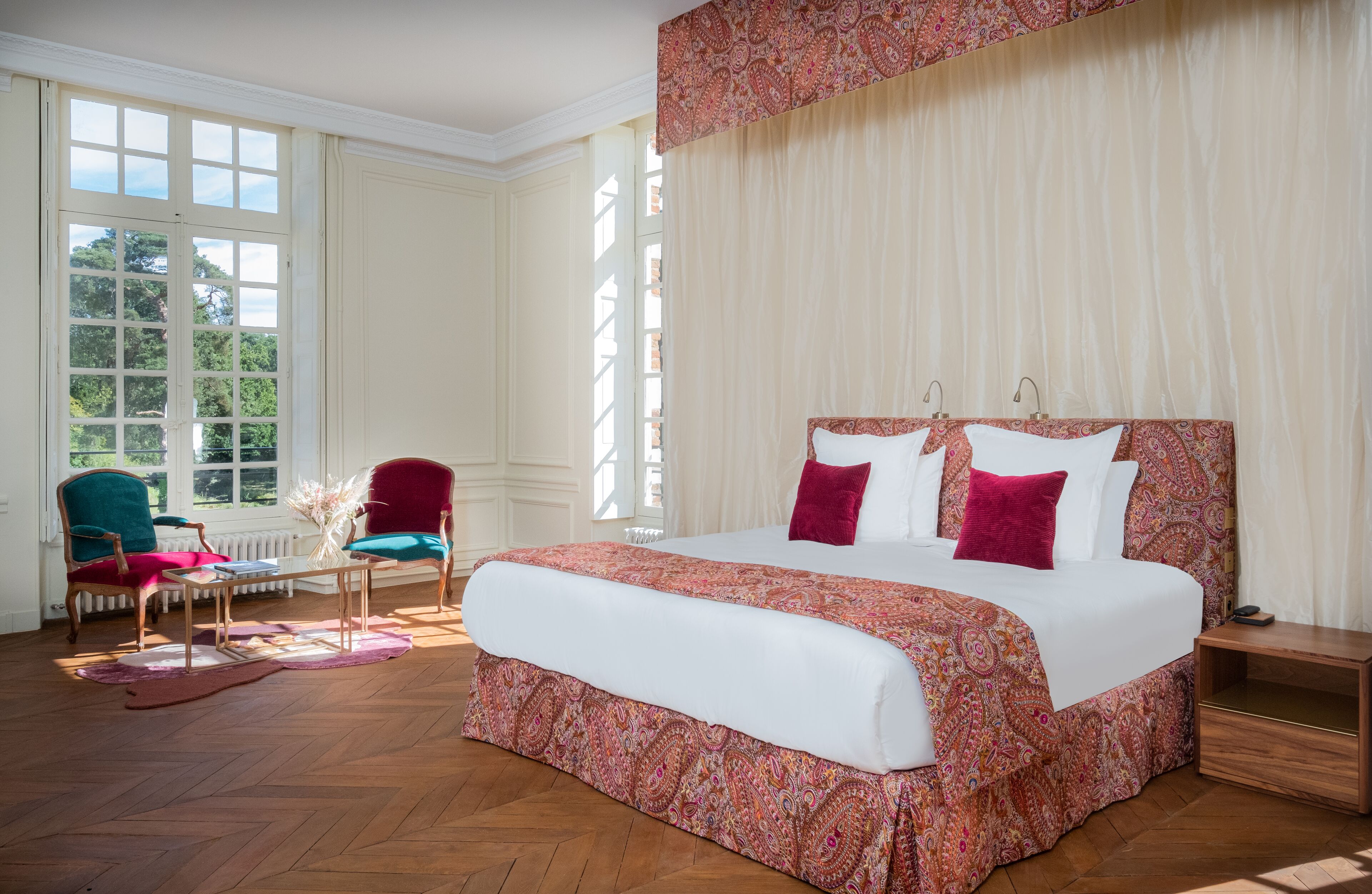Un dormitorio espacioso con una cama grande, una zona de estar con sillas y vistas a zonas verdes por las ventanas.