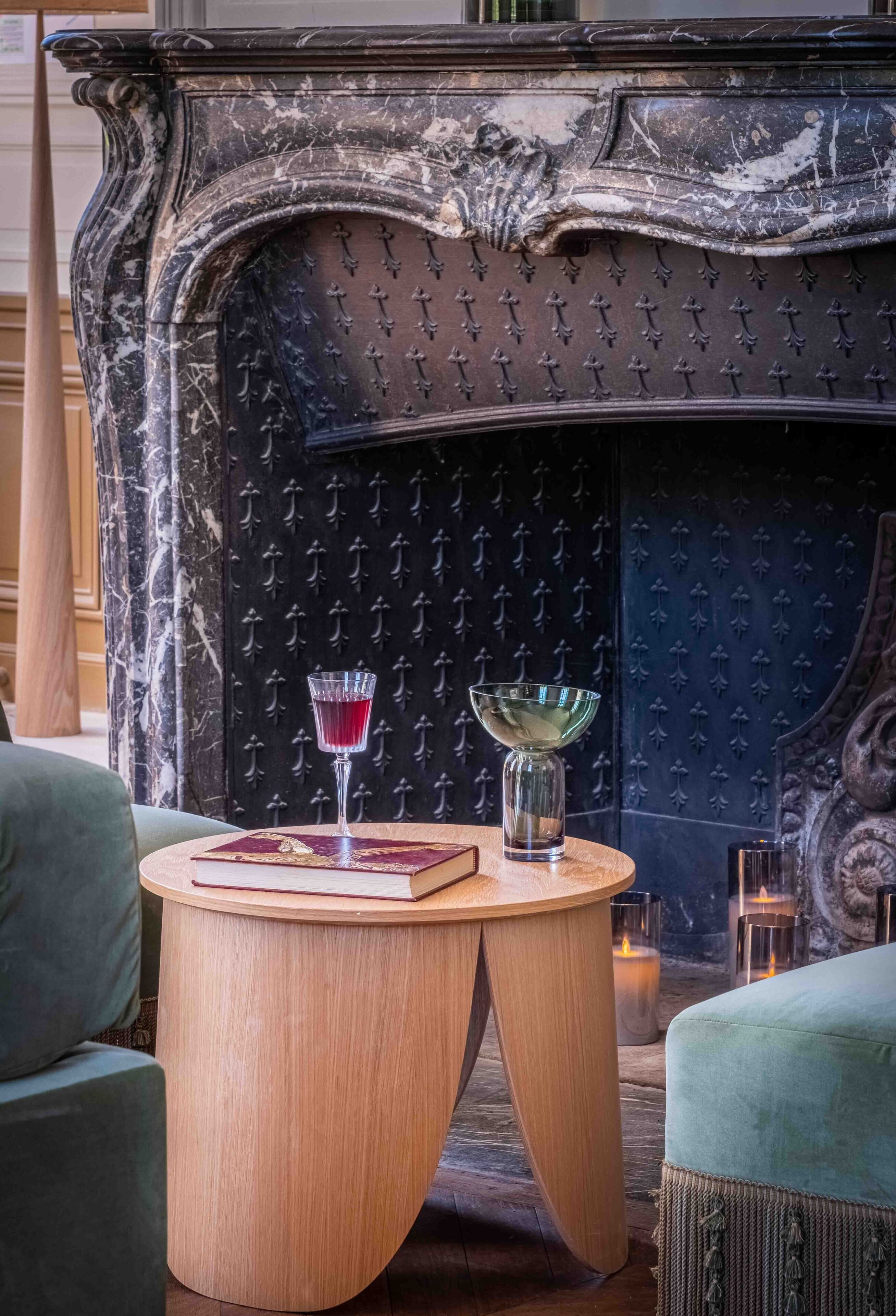 Une cheminée aux motifs complexes, une table ronde en bois sur laquelle sont posés un livre et un verre, et deux fauteuils verts recouverts de tissu.