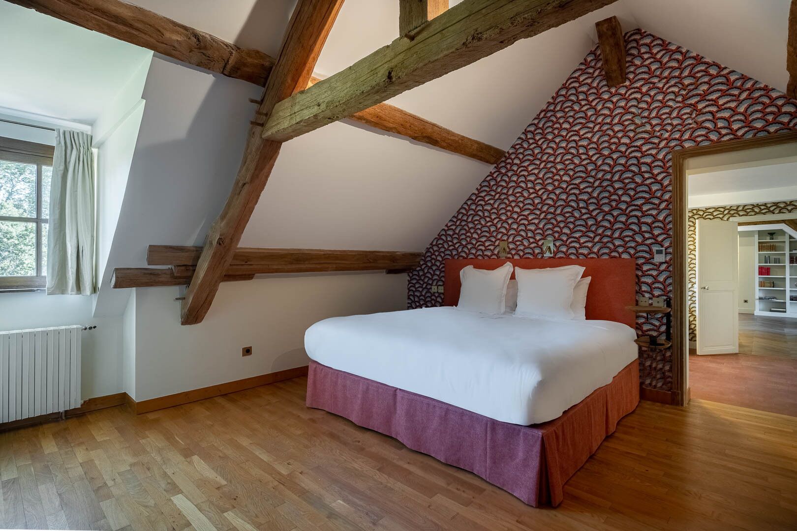 A bedroom with a sloped ceiling, wooden beams, a bed with white bedding, a patterned wall, and a view of a library.