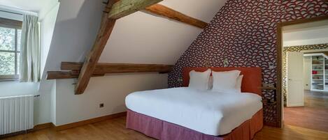A bedroom with a sloped ceiling, wooden beams, a bed with white bedding, a patterned wall, and a view of a library.
