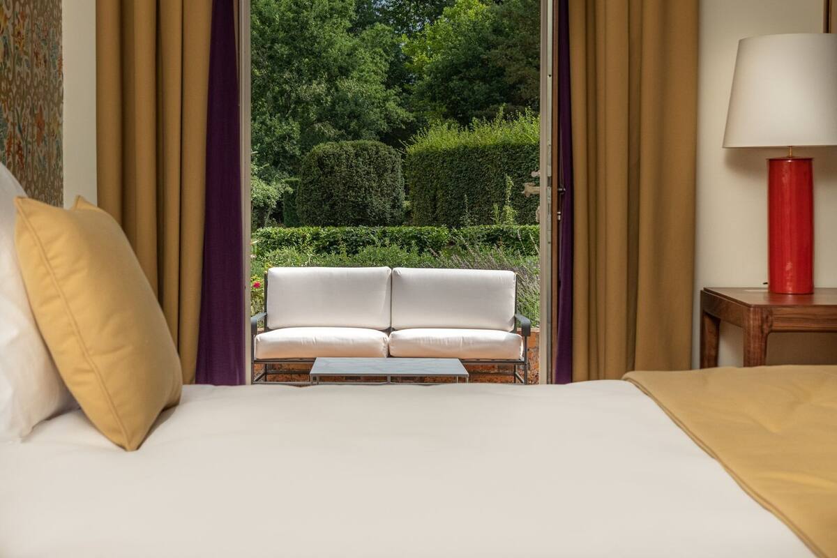Een hotelkamer met een bed, een witte bank, een rode lamp en uitzicht op een tuin door een geopende deur.