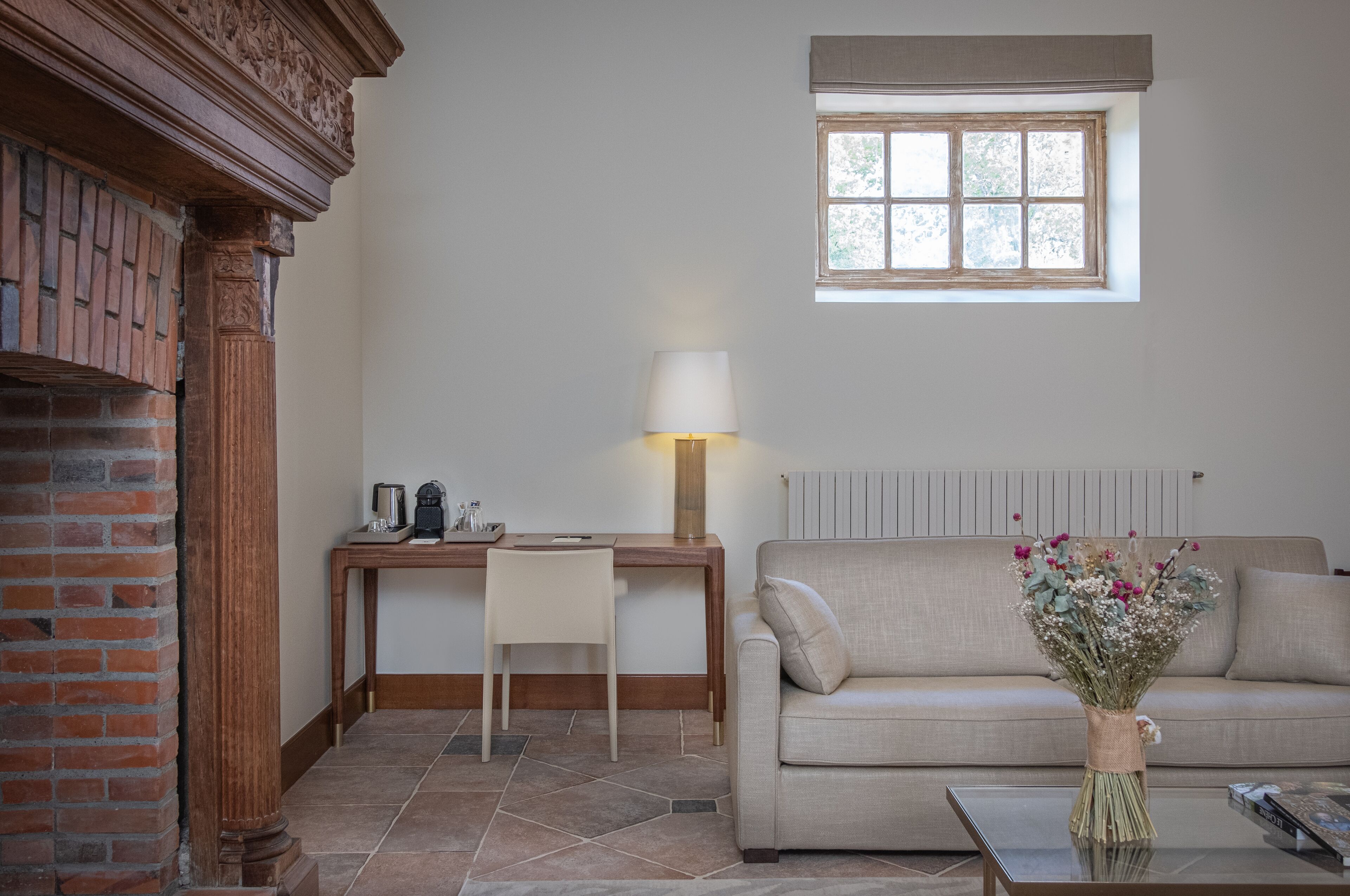 Een woonkamer met een open haard van baksteen, een houten bureau met een lamp, een beige bank en een vaas met bloemen op een bijzettafeltje.