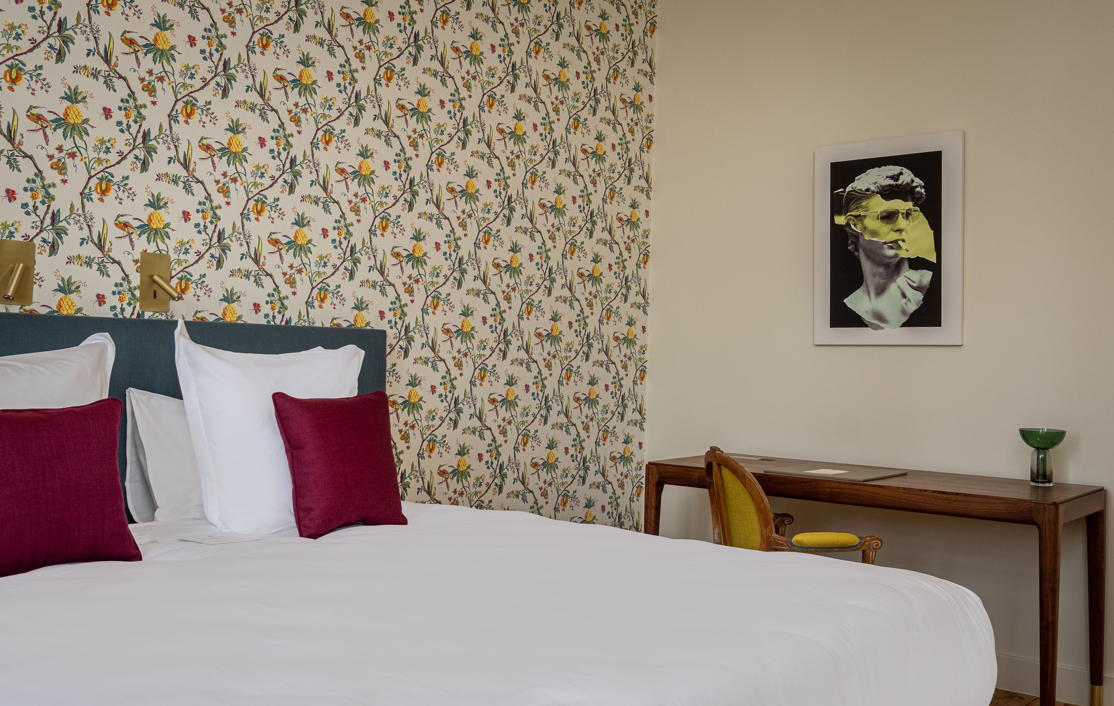 A neatly made bed with white and red pillows, a floral wallpaper, a wooden nightstand with a green bowl, and a framed artwork on the wall.