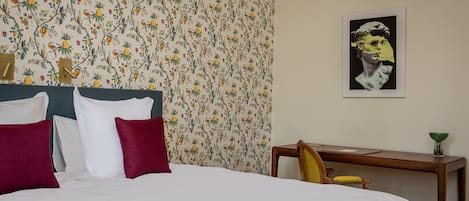 A neatly made bed with white and red pillows, a floral wallpaper, a wooden nightstand with a green bowl, and a framed artwork on the wall.