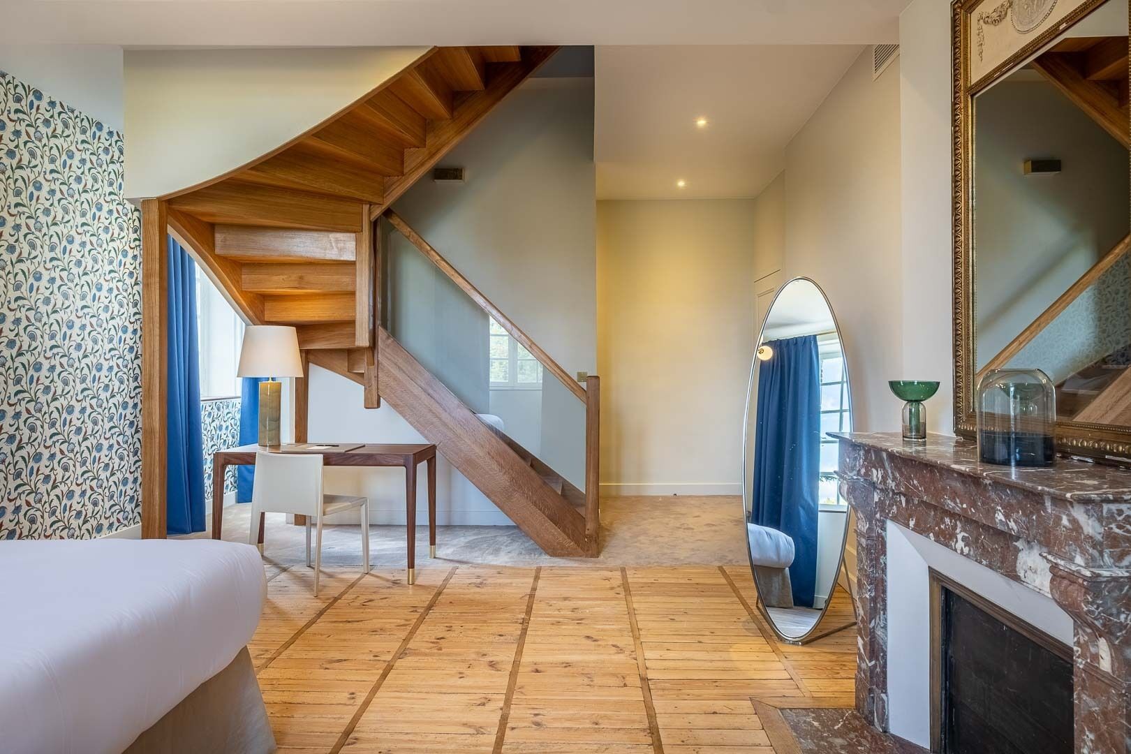 Ein Zimmer mit einer Holztreppe, einem Bett, einem Schreibtisch mit Lampe, einem Spiegel und einem Kamin.