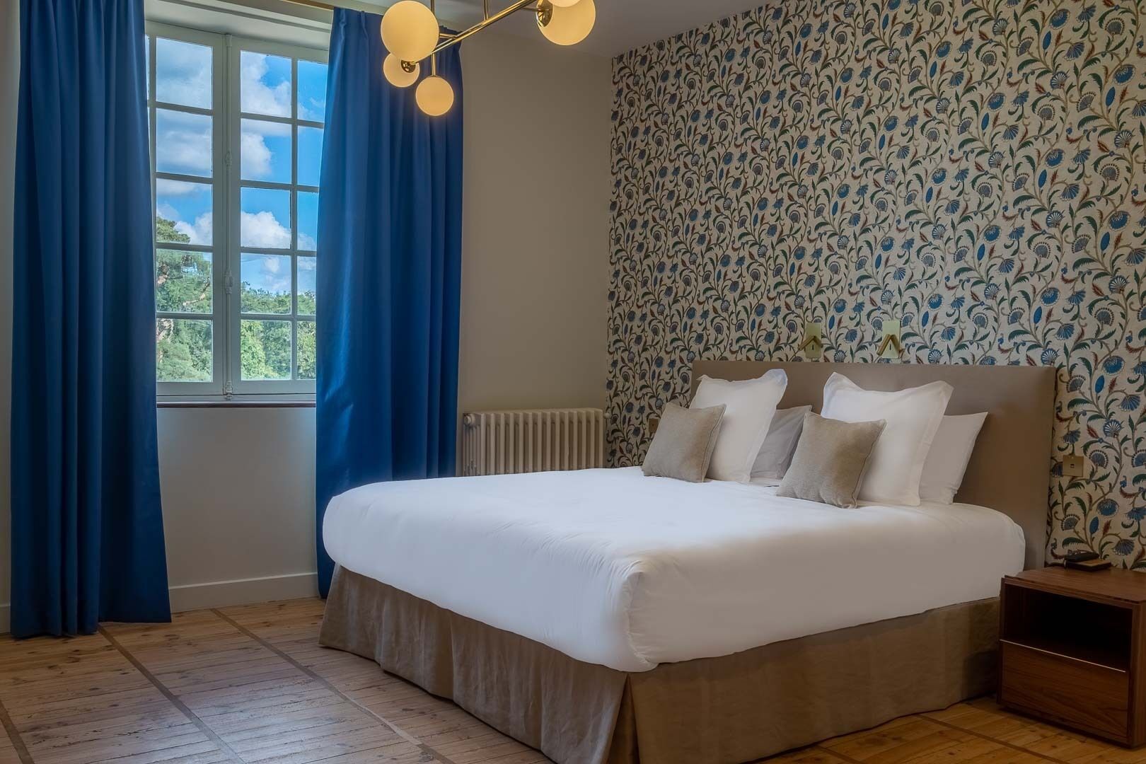 A bedroom with a large bed, blue curtains, a patterned wallpaper, and a wooden floor.