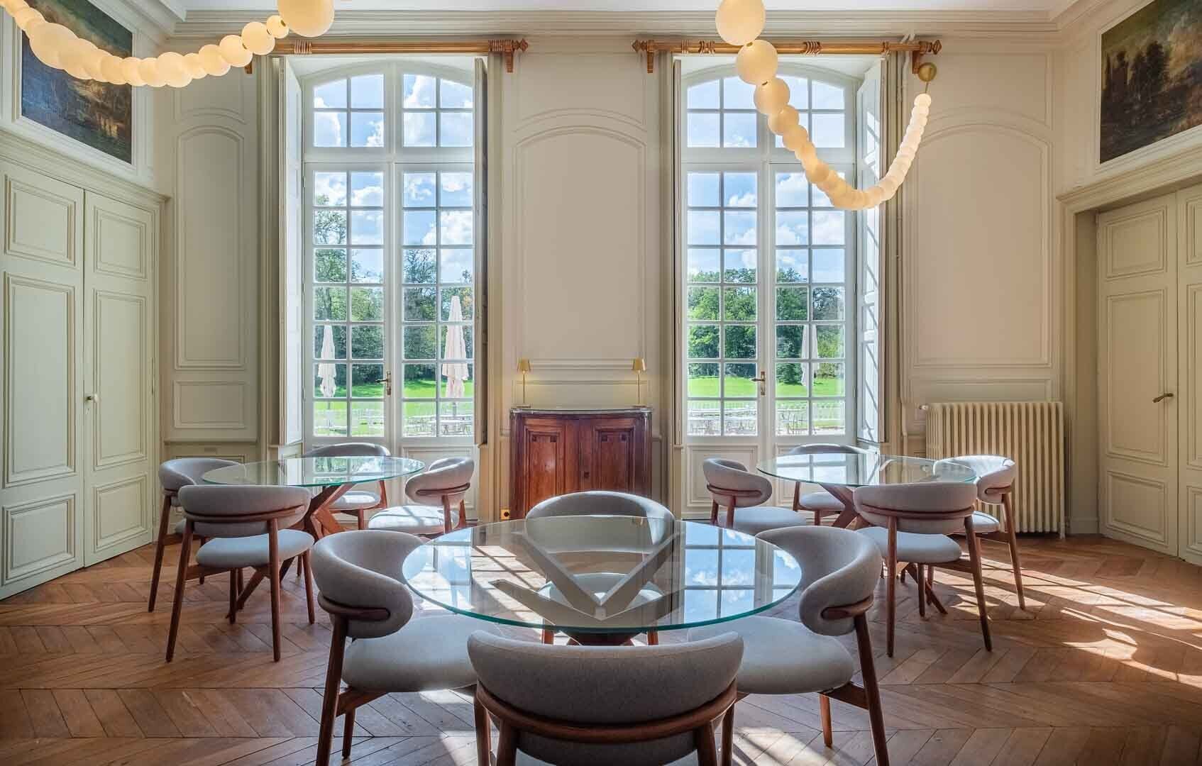 A dining room with a round glass table surrounded by six chairs, large windows with a view of greenery, and a classic architectural style.