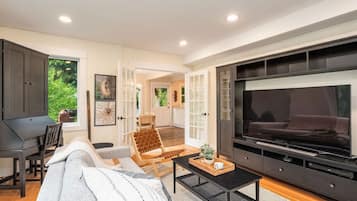 A modern living room with a large flat-screen TV, a sofa, a coffee table, and a dining area with a piano.