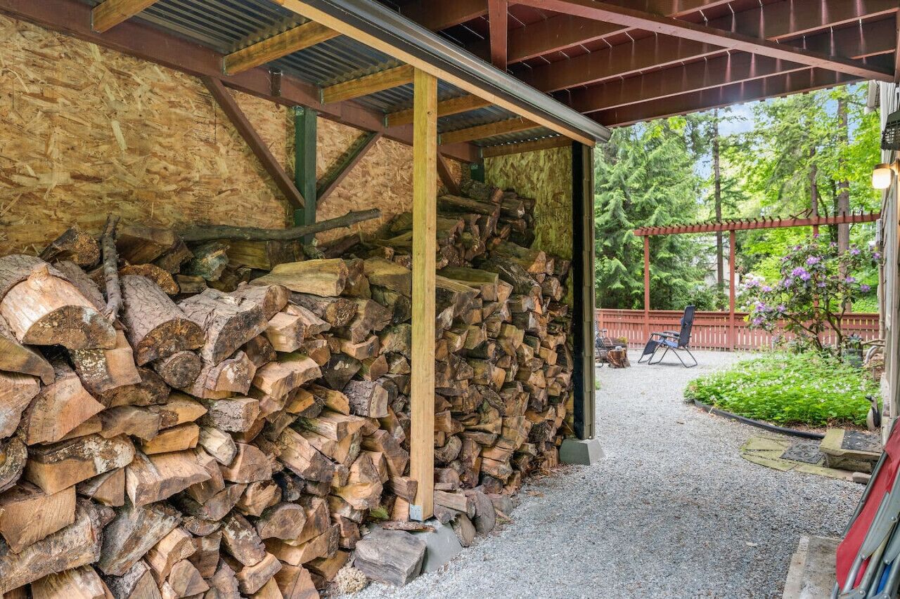 Un espace extérieur abrité comprenant une pile de bois de chauffage, une pergola et un jardin agrémenté de plantes et d’une chaise.