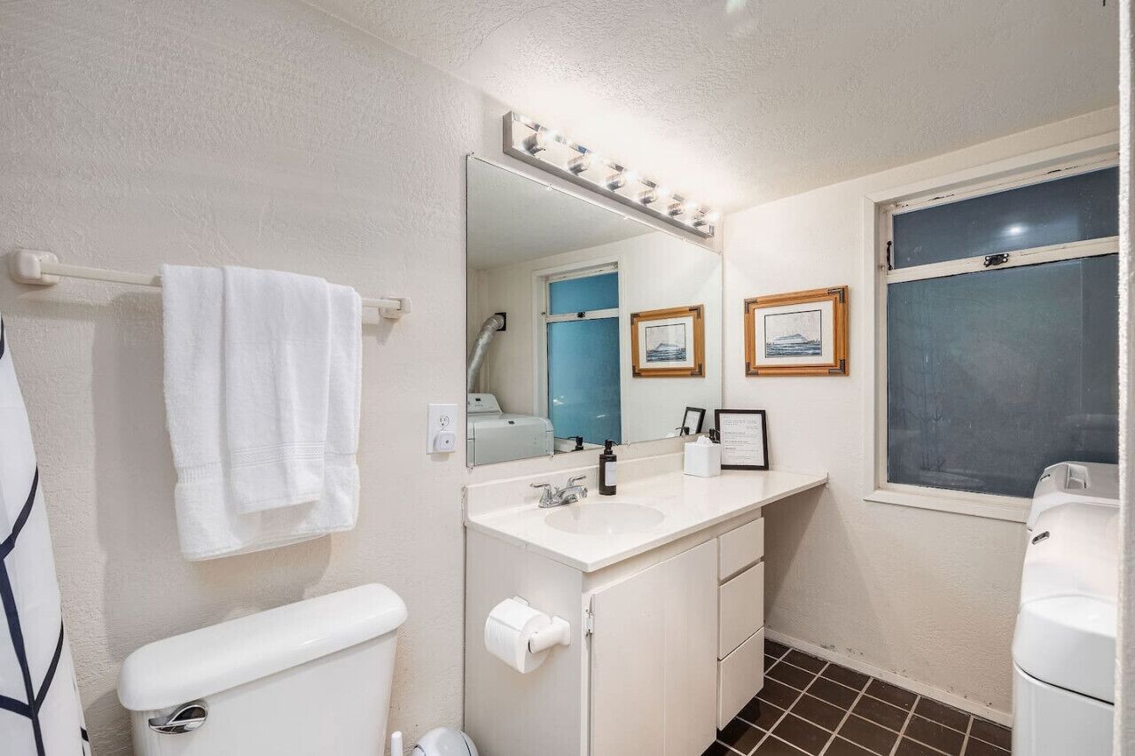 Een badkamer met toilet, wastafel, opbergruimte en een grote spiegel met verlichting. Twee ingelijste foto's hangen aan de muur en er is een raam met uitzicht.